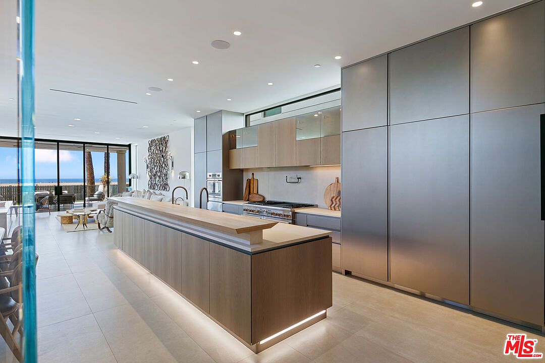 This modern, open-concept kitchen features a large island with a light wood base and a stone countertop, complemented by sleek, metallic-finish cabinetry. The space is illuminated by recessed lighting and under-cabinet lighting, creating a warm and sophisticated atmosphere. A wide opening leads to a bright living area with large glass doors that provide a clear view of the ocean beyond.