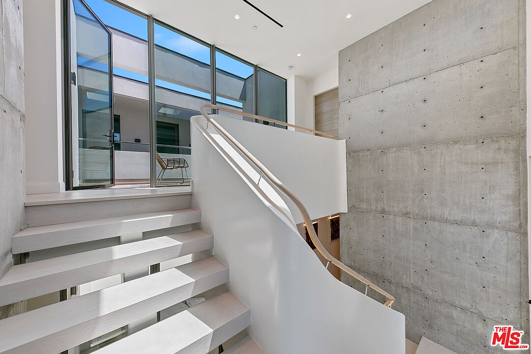 This modern interior space features a striking staircase with light wood treads and a sleek white railing that curves gracefully upward. To the right, a large, raw concrete wall adds an industrial texture, while to the left, expansive glass doors open onto an outdoor balcony area. The perspective captures the clean lines and minimalist aesthetic of the home, emphasizing the bright, airy atmosphere created by the natural light.
