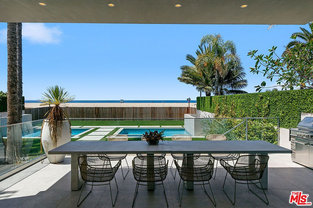 This covered outdoor patio features a long, modern dining table with wire-frame chairs, overlooking a private swimming pool and a lush green lawn. Beyond the yard, a wooden fence provides privacy before opening up to a clear view of the beach and ocean horizon. The space is designed for seamless indoor-outdoor living, with recessed lighting in the ceiling and a clean, minimalist aesthetic.