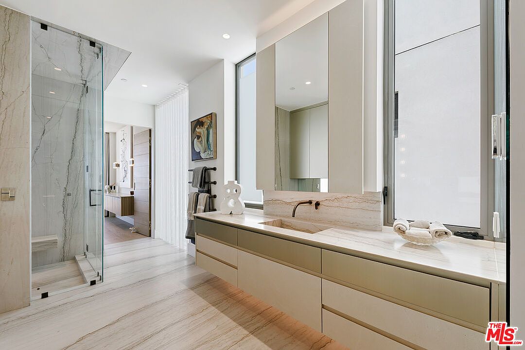 This modern primary bathroom features a sleek, floating vanity with a stone countertop and integrated sink, complemented by a large, wall-mounted mirror. To the left, a glass-enclosed walk-in shower showcases light-toned, veined stone wall tiles that match the flooring. The space is brightened by tall windows that allow natural light to fill the room, creating a clean and luxurious atmosphere.