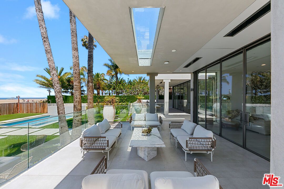 This expansive covered patio offers a seamless transition between indoor and outdoor living, featuring a modern seating arrangement with light-toned sofas and a central stone-topped coffee table. The space is defined by a sleek, minimalist aesthetic, with large glass sliding doors providing a clear view into the home's interior. Beyond the glass railing, a well-manicured lawn, a swimming pool, and tall palm trees create a serene, resort-like atmosphere overlooking the nearby beach.
