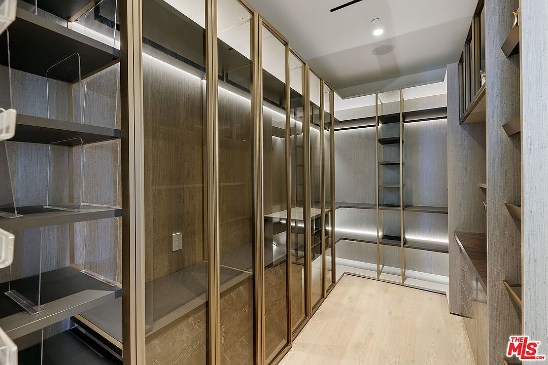 This sophisticated walk-in closet features custom cabinetry with a warm wood finish and integrated LED lighting. The space is defined by floor-to-ceiling glass-fronted wardrobe doors with metallic frames, creating a sleek and organized aesthetic. Light-toned wood flooring runs throughout the room, enhancing the bright and luxurious feel of this high-end storage area.