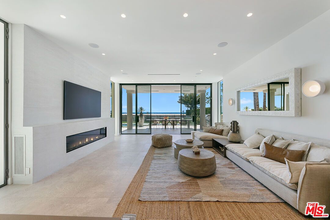 This bright, contemporary living room features a neutral color palette and a seamless transition to an outdoor patio through large glass sliding doors. The space is anchored by a long, linear fireplace set into a light-toned wall and a large area rug layered over light stone flooring. A comfortable sofa and circular coffee tables create an inviting seating area, while a large mirror on the wall enhances the sense of depth and natural light.