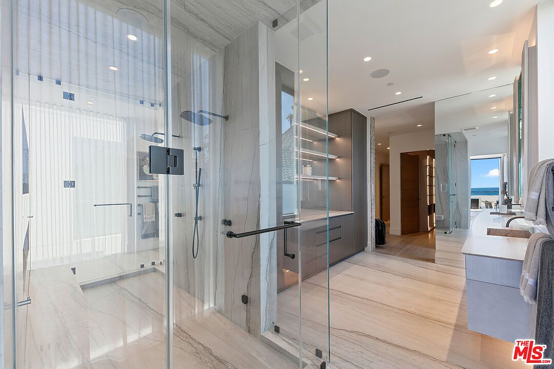 This luxurious primary bathroom features a spacious walk-in shower enclosed by floor-to-ceiling glass panels, showcasing light-toned stone walls and flooring with subtle veining. To the right, a sleek vanity area includes built-in cabinetry with open shelving and integrated lighting, leading toward a bright space with a view of the ocean. The perspective is captured from the shower entrance, emphasizing the open, airy layout and high-end contemporary design.