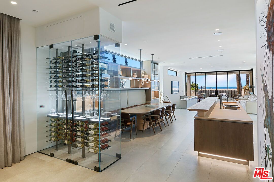 This open-concept living space features a sleek, modern kitchen with a large island and a prominent glass-enclosed wine display. A long dining table sits adjacent to the kitchen, leading the eye toward large glass doors that open to a scenic beach view. The interior design is characterized by a neutral color palette, clean lines, and a seamless flow between the culinary and dining areas.