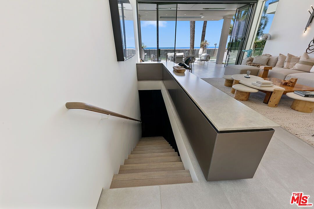 This perspective captures a modern staircase descending into a dark lower level, positioned adjacent to a bright, open-concept living area. The staircase features light wood treads and a sleek handrail, while a long, stone-topped console serves as a partition between the stairs and the living space. Beyond the living area, large glass doors open to reveal a scenic coastal view, emphasizing the home's seamless indoor-outdoor connection.