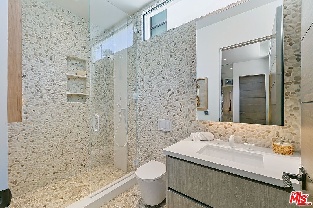 This modern bathroom features a distinctive pebble-tiled wall that extends into the walk-in shower, creating a natural, textured aesthetic. The space includes a floating vanity with a white countertop, a wall-mounted toilet, and a large, frameless glass shower enclosure. A tall, rectangular mirror reflects the room, while a clerestory window above the shower allows for natural light to brighten the area.