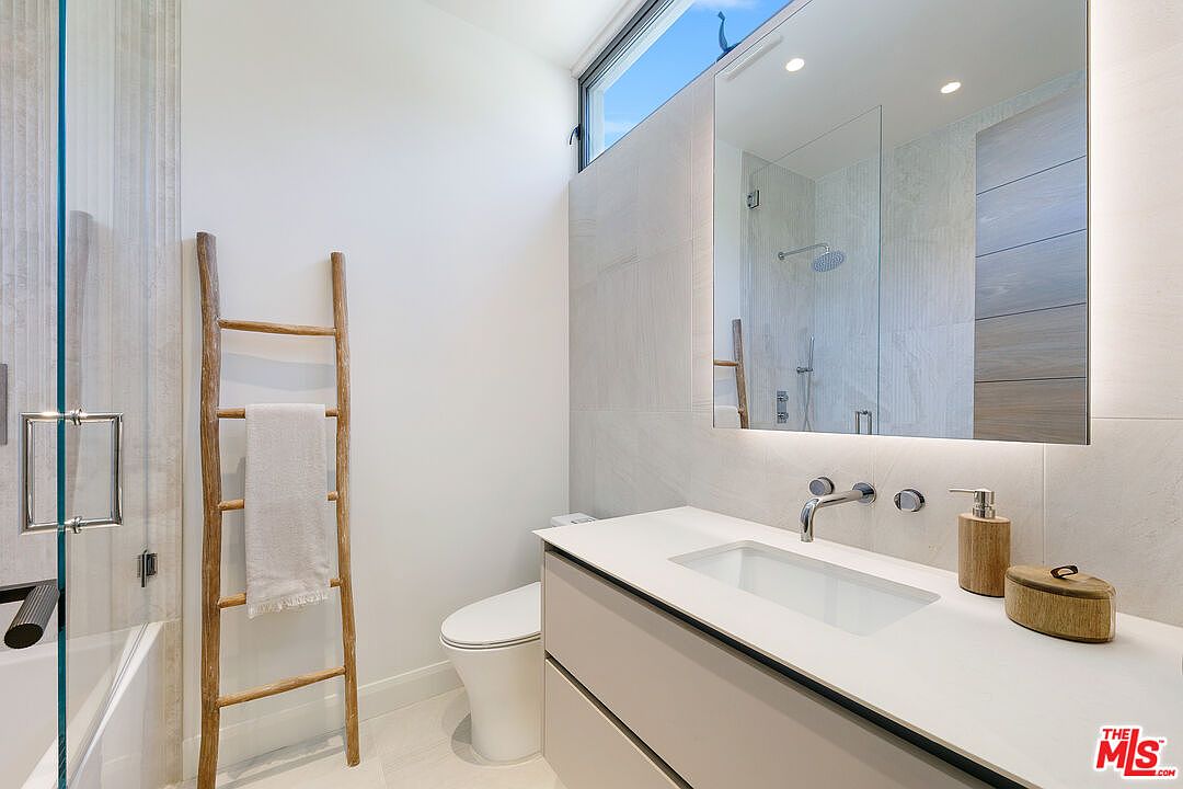 This modern, minimalist bathroom features a sleek vanity with a white countertop and a large, frameless mirror reflecting the shower area. A decorative wooden ladder serves as a towel rack against the crisp white wall, adding a touch of organic texture to the space. The room is brightened by a clerestory window that allows natural light to filter in, highlighting the clean lines and neutral color palette.