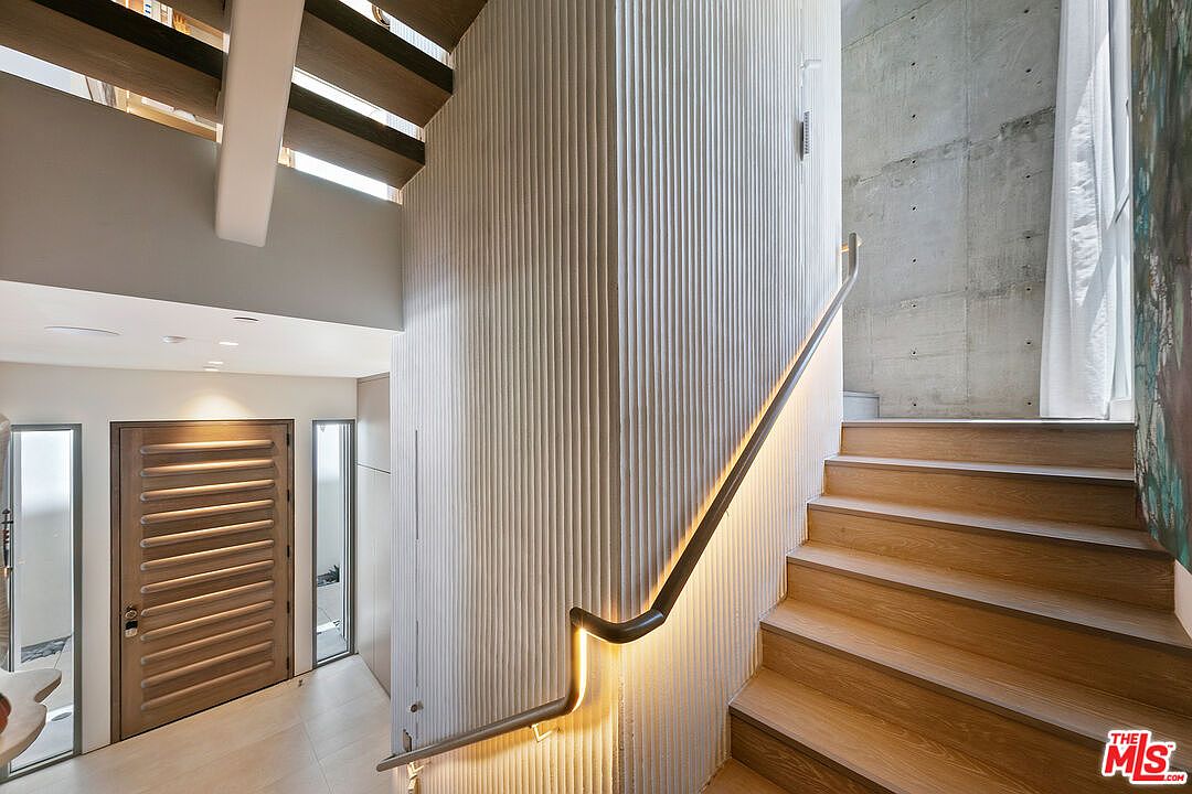This modern entryway features a striking staircase with light wood treads and a sleek, illuminated handrail that follows the curve of a textured, vertical-slatted wall. To the left, a contemporary front door with horizontal panels is flanked by tall, narrow windows, while a raw concrete wall adds an industrial touch to the stairwell. The perspective captures the transition from the entry level to the upper floor, highlighting the sophisticated blend of natural textures and architectural lighting.