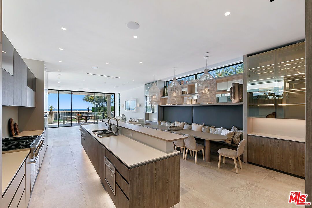This expansive, modern kitchen features a long central island with a built-in sink and integrated appliances, paired with a custom banquette dining area along the wall. The space is characterized by clean lines, light-toned cabinetry, and a seamless transition to an outdoor patio visible through large sliding glass doors. Three woven pendant lights hang above the dining table, creating a warm, inviting atmosphere that emphasizes an open-concept layout.