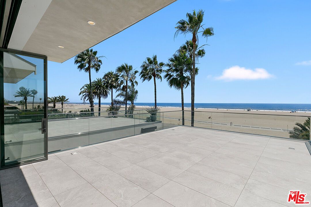 This expansive, modern balcony features large-format light-toned tiles and a sleek glass railing that provides an unobstructed view of the beach and ocean. The space is partially covered by a deep overhang, creating a seamless transition from the interior through large sliding glass doors. The scene captures a bright, sunny day with palm trees framing the coastal horizon, emphasizing a luxurious and serene outdoor living experience.