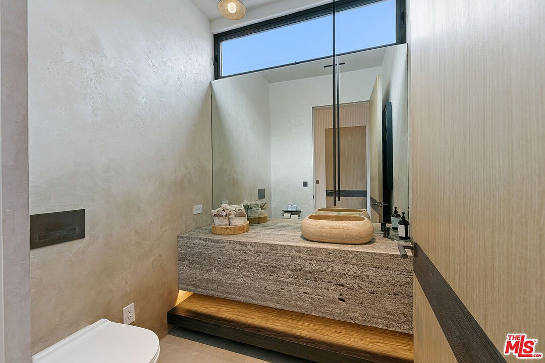 This modern powder room features a floating vanity crafted from textured stone, topped with a unique, rounded stone vessel sink. A large, frameless mirror reflects the space, while a tall, narrow window above provides natural light. The room is finished with textured, neutral-toned walls and a warm wood-toned base, creating a sophisticated and minimalist aesthetic.