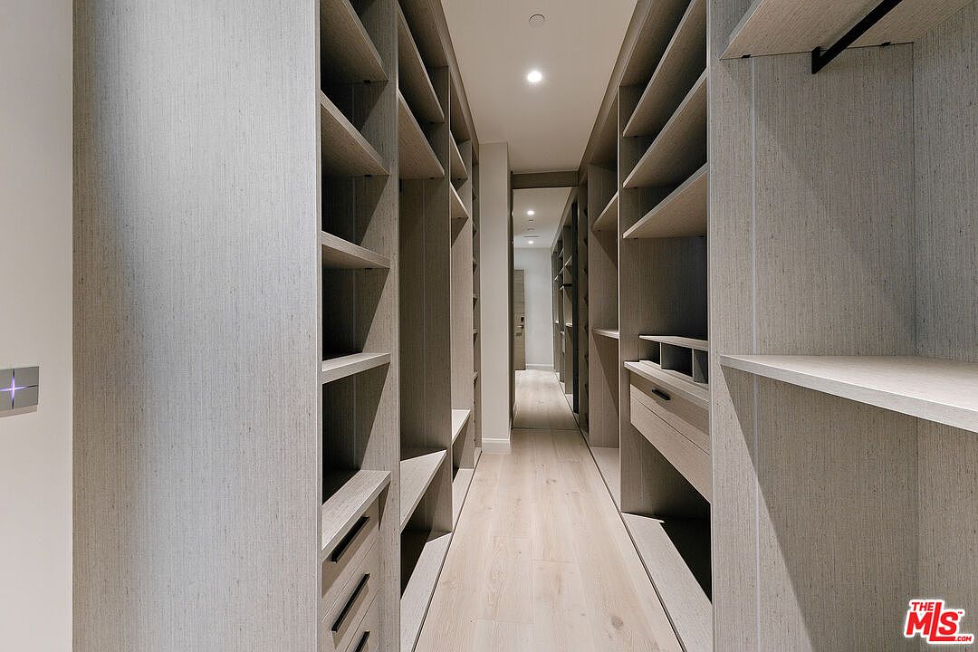 This spacious walk-in closet features extensive custom light wood shelving and cabinetry that spans both sides of the narrow, elongated room. The design includes a mix of open shelving, hanging rods, and integrated drawers, all finished in a clean, contemporary aesthetic. A central recessed light illuminates the light-toned wood flooring, creating a bright and organized atmosphere.