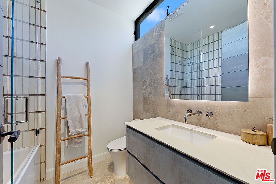 This modern bathroom features a sleek vanity with a light-toned countertop and a large, rectangular mirror reflecting the shower area. A decorative wooden ladder towel rack adds a rustic touch against the neutral-toned wall tiles, while a tall, narrow window provides natural light. The space is finished with a minimalist aesthetic, including a wall-mounted toilet and clean, contemporary fixtures.
