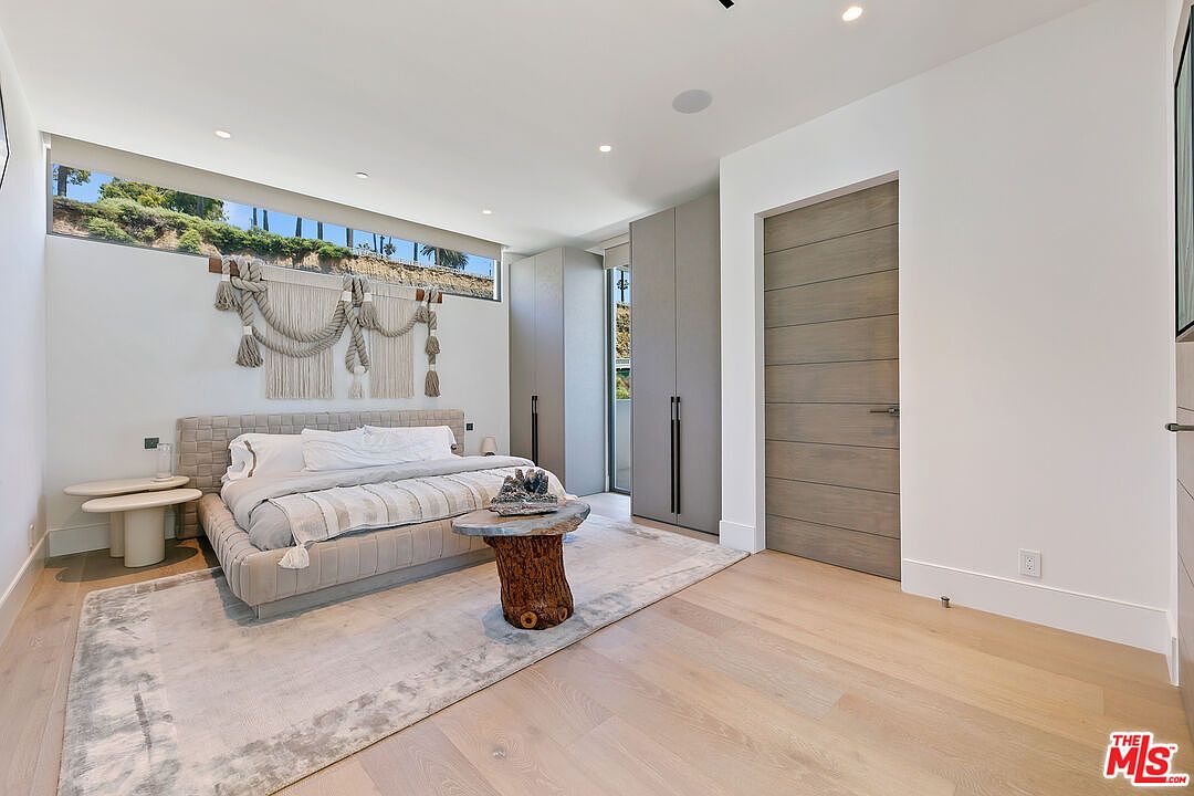 This modern primary bedroom features a low-profile upholstered bed centered against a wall adorned with a decorative macramé wall hanging. A unique, natural-edge wood stump serves as a coffee table, while light wood flooring and neutral-toned cabinetry provide a clean, contemporary aesthetic. A long, horizontal window above the bed allows natural light to filter into the space, creating an airy and serene atmosphere.