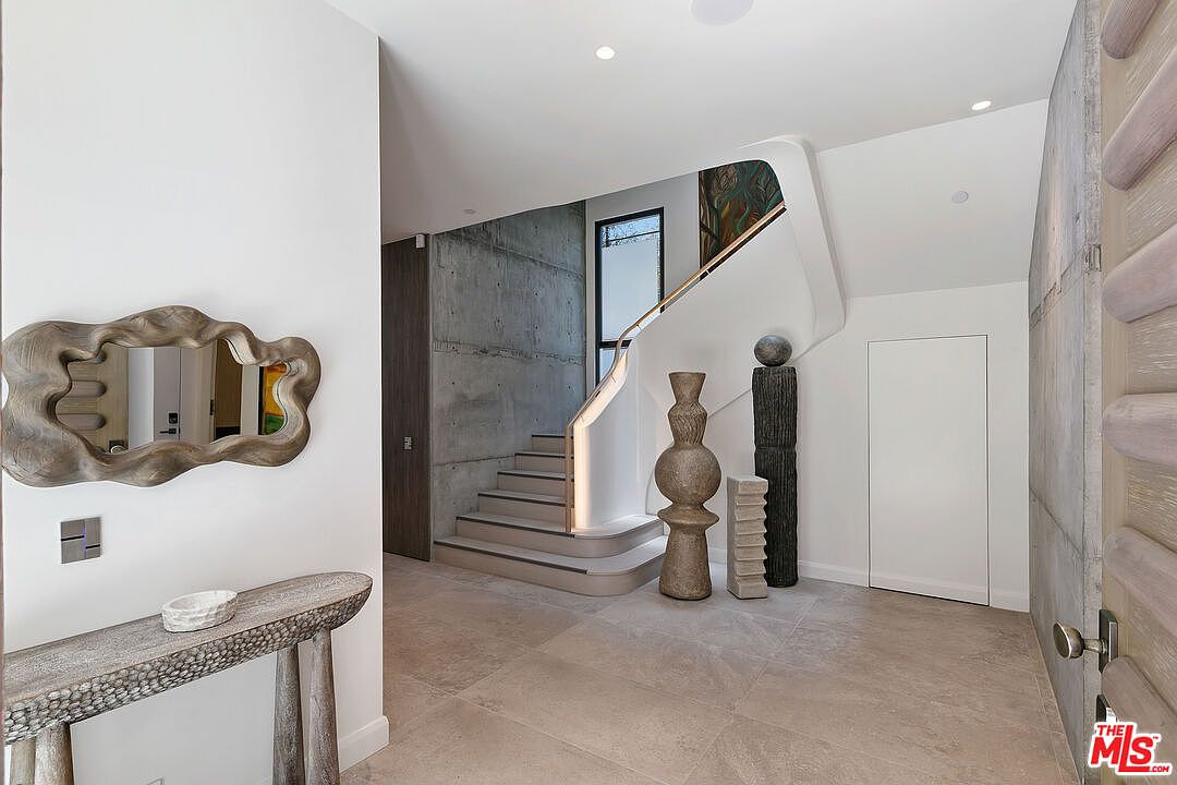 This modern entryway features a striking staircase with a curved, illuminated handrail set against a raw concrete wall. The space is decorated with sculptural art pieces and a textured, organic-shaped mirror above a console table, creating a sophisticated and artistic atmosphere. The neutral color palette and clean lines emphasize a minimalist, high-end architectural design.