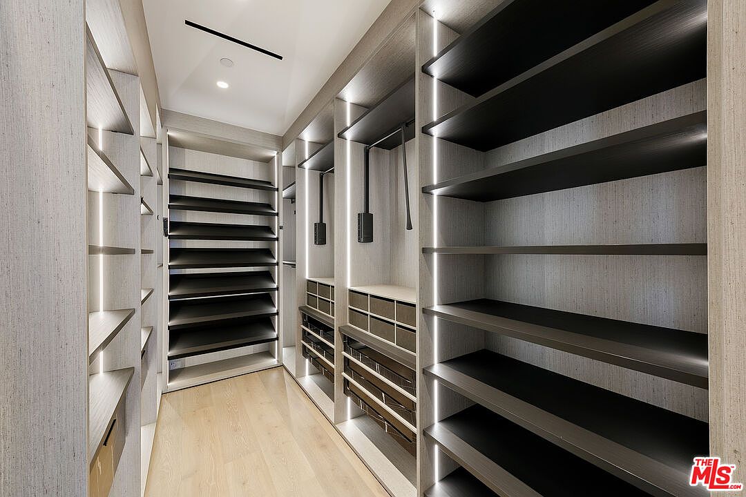 This modern walk-in closet features custom light wood cabinetry with integrated vertical LED lighting strips that illuminate the shelving and hanging sections. The space includes a mix of open shelving, angled shoe racks, and built-in drawers, all set against a light wood floor. The clean, minimalist design creates a sophisticated and organized atmosphere for storage.