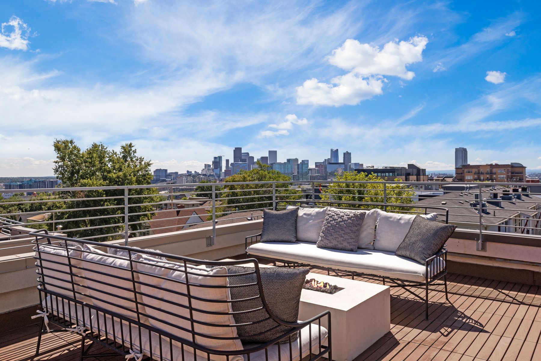 This image showcases a stylish rooftop deck with comfortable seating arrangements, including sofas and a modern fire pit. The deck features wooden flooring and a sleek railing, offering an unobstructed view of the city skyline under a bright, partly cloudy sky. The scene evokes a sense of luxury and relaxation, perfect for outdoor entertaining.