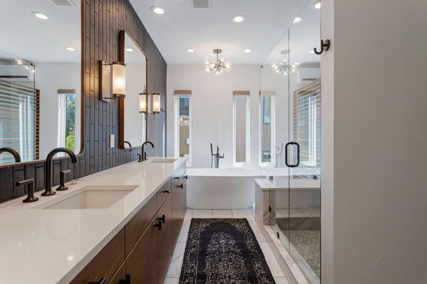This is a well-lit primary bathroom featuring a double vanity with a white countertop and dark wood cabinets. A large mirror and modern sconces adorn the wall above the vanity. A freestanding bathtub is positioned between two narrow windows, and a glass-enclosed shower is visible to the right, creating a luxurious and spa-like atmosphere.