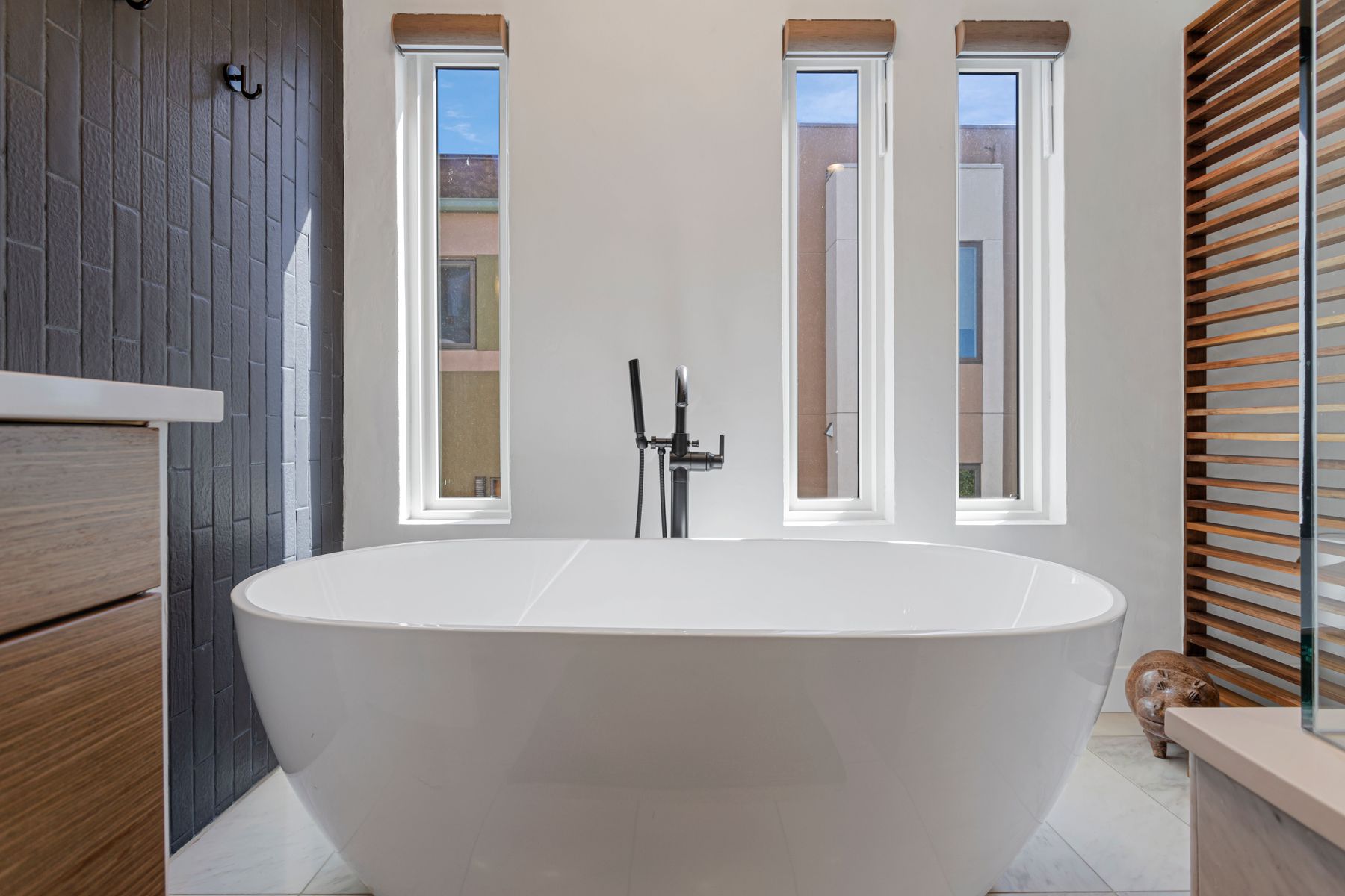This is a bright and modern primary bathroom featuring a freestanding white bathtub as the focal point. Three tall, narrow windows provide natural light and views, while a dark-tiled wall adds a touch of contrast. The room is clean and minimalist, creating a spa-like atmosphere.