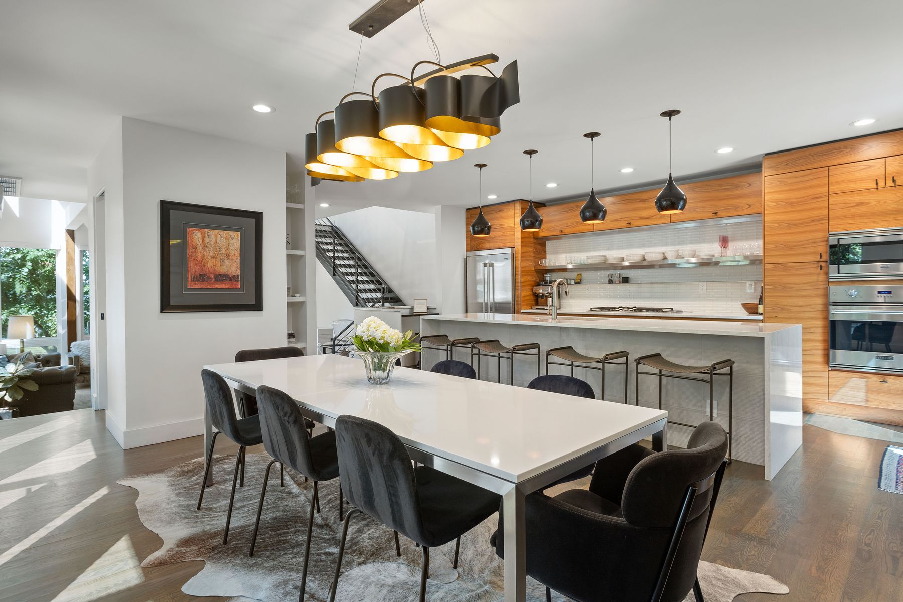 This interior shot showcases a modern dining area adjacent to a kitchen. The dining room features a white table with black velvet chairs, set upon a cowhide rug. A contemporary chandelier hangs above the table, and the open floor plan leads into a sleek kitchen with wood cabinetry and a breakfast bar, creating a seamless and stylish living space.
