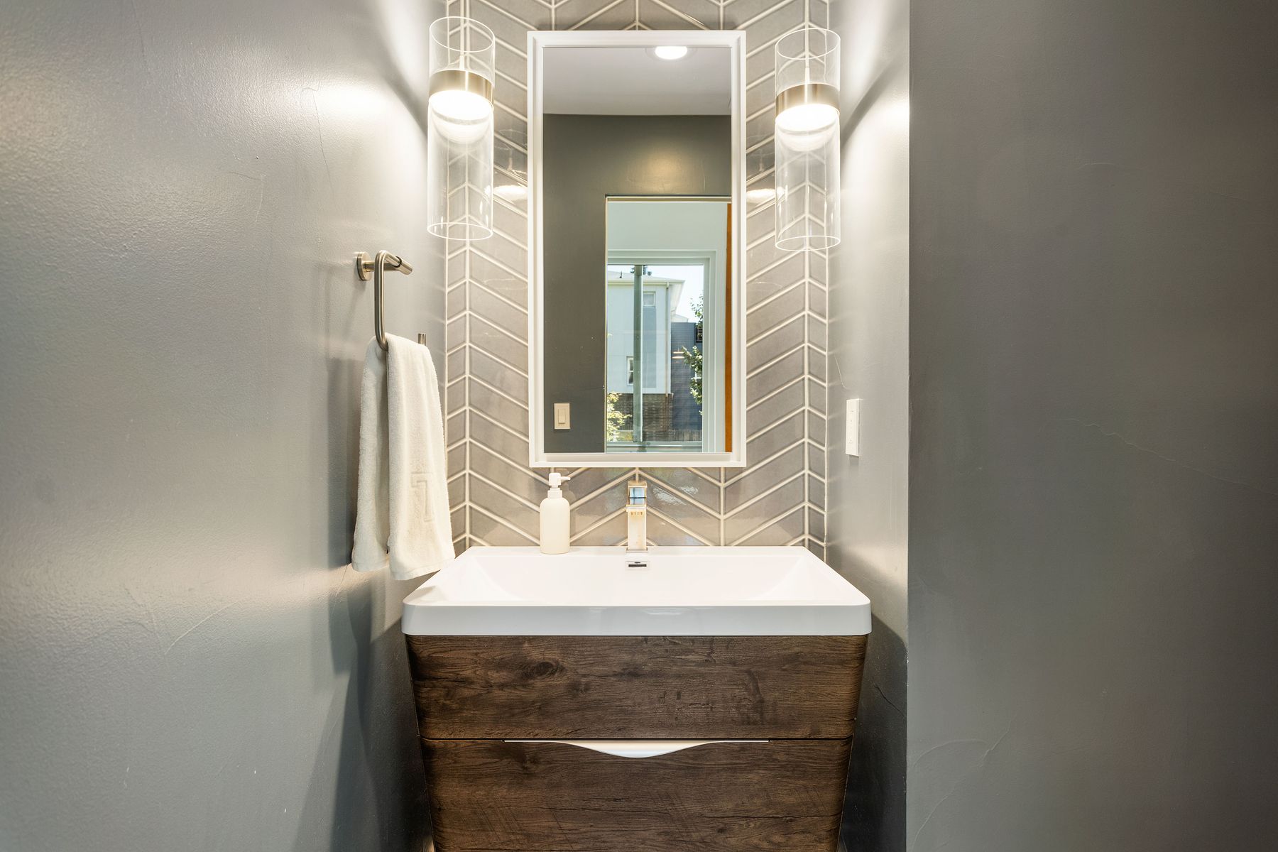 This is a well-lit bathroom featuring a modern vanity with a white countertop and a wood-grain cabinet. A rectangular mirror is mounted above the sink, flanked by stylish sconce lighting. The walls are painted in a neutral gray tone, and the backsplash behind the vanity has a herringbone pattern, adding a touch of visual interest.