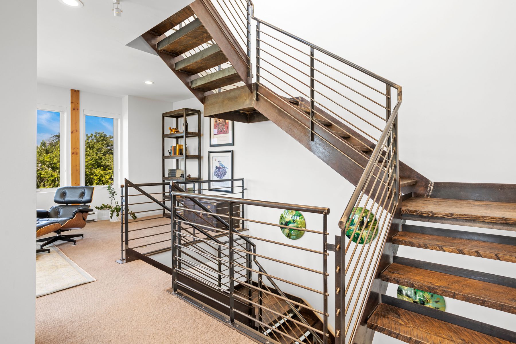 This interior shot showcases a modern staircase with a unique industrial design. The stairs feature a combination of wood and metal, with metal railings and stringers adding an edgy, contemporary feel. The surrounding walls are white, providing a clean backdrop that highlights the staircase's architectural details and the natural light coming from the windows.
