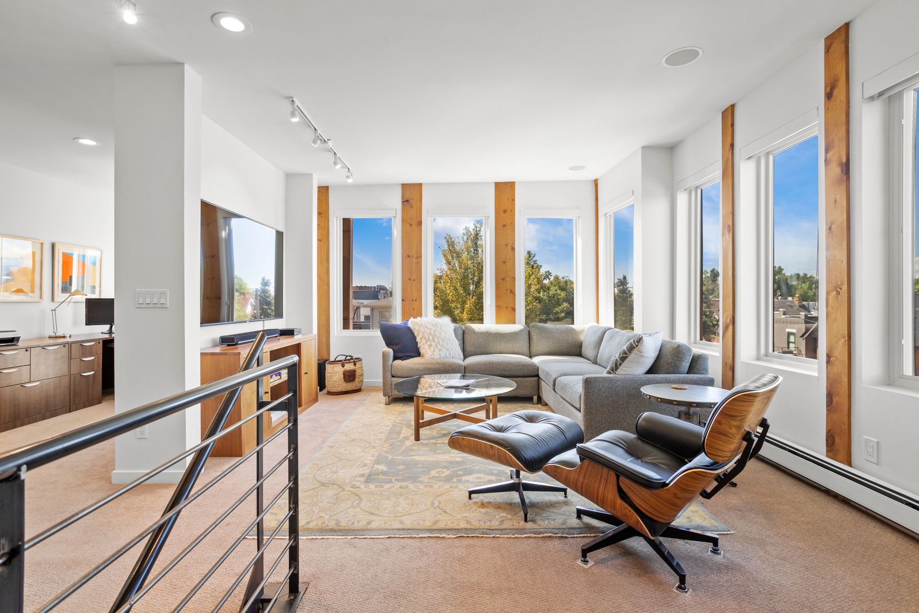This is a bright and airy living room featuring a large sectional sofa, a modern chair with ottoman, and a glass-topped coffee table. The room is well-lit with natural light streaming through multiple windows framed by wooden accents. The space is decorated in neutral tones with a patterned area rug, creating a comfortable and inviting atmosphere.