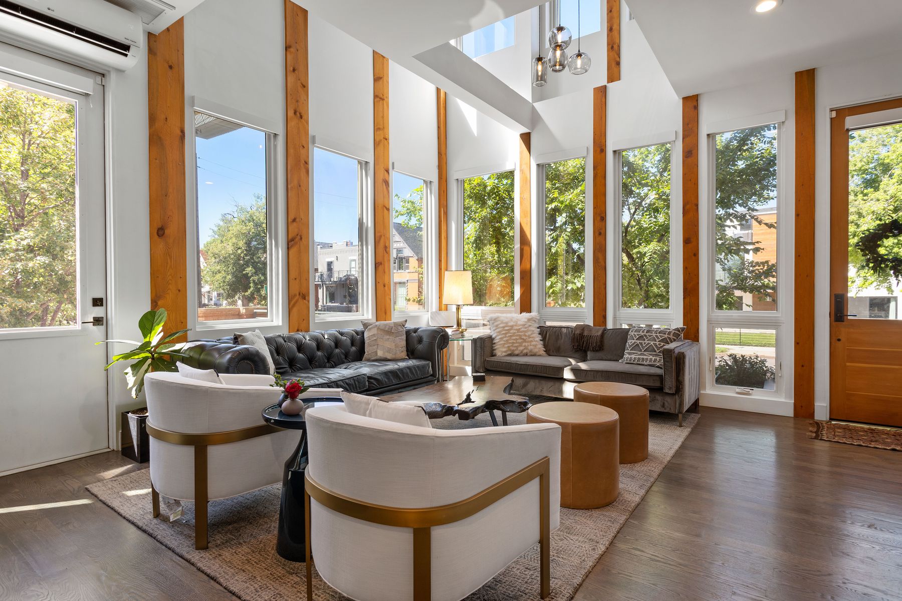 This is a bright and airy living room featuring a high ceiling with skylights and large windows that offer ample natural light. The room is furnished with a mix of modern and classic pieces, including a tufted leather sofa, a gray sofa, and two white armchairs with gold accents. The hardwood floors and neutral-toned rug add warmth and texture to the space.