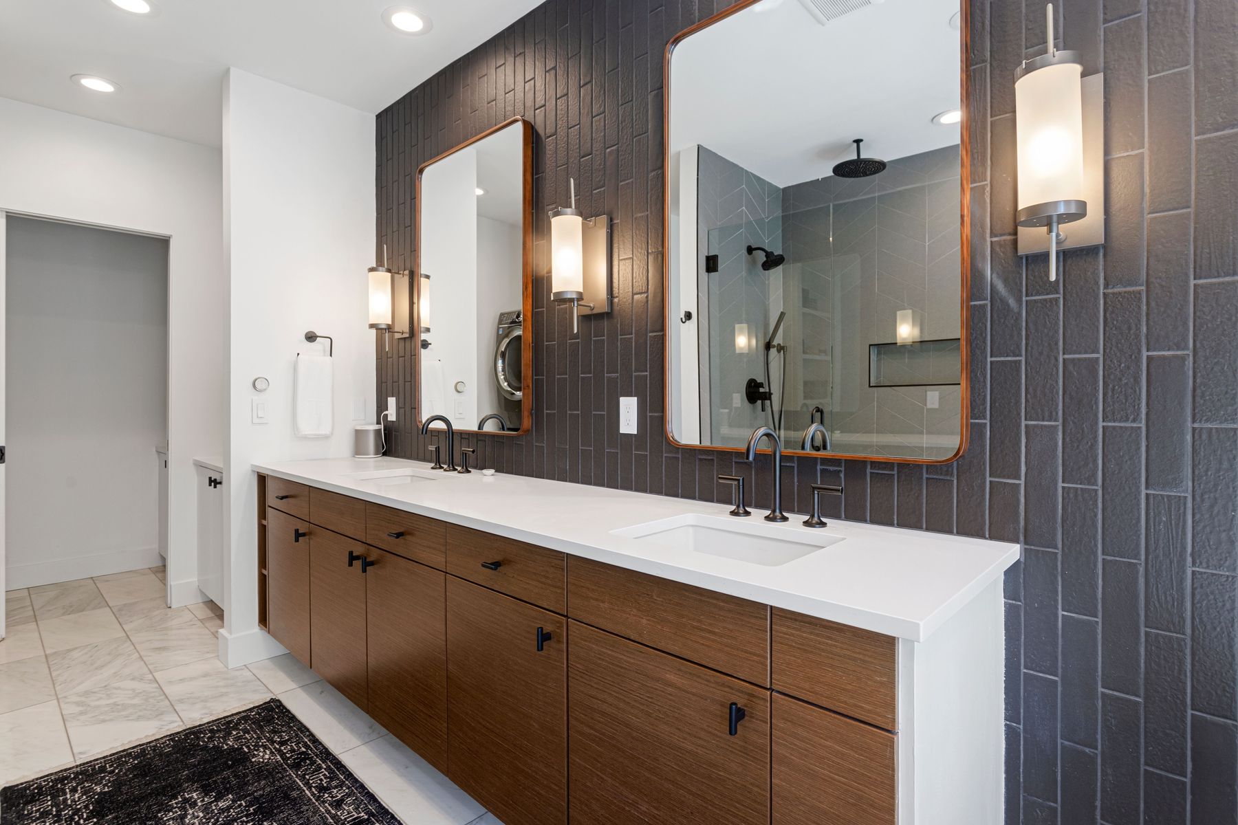 This is a primary bathroom featuring a double vanity with a white countertop and wood-grain cabinets. Two rectangular mirrors with copper frames are mounted above the sinks, flanked by modern sconces. The wall behind the vanity is covered in dark gray, vertically stacked tiles, and a glimpse of a glass-enclosed shower can be seen in one of the mirrors.