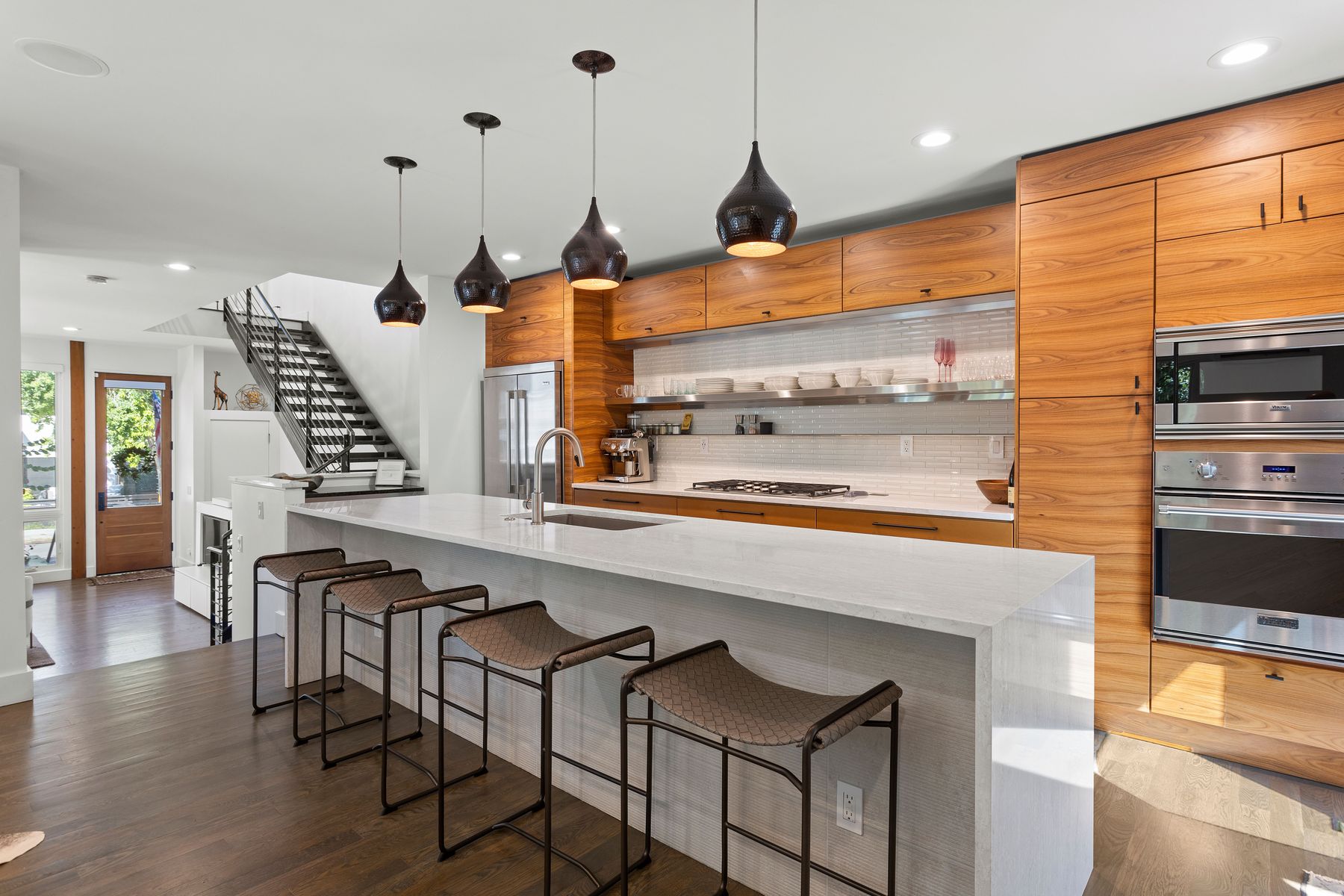 This is a well-lit, modern kitchen featuring sleek wooden cabinetry and stainless steel appliances, including a built-in oven and microwave. A large white countertop island with seating anchors the space, illuminated by pendant lights. The view includes a staircase and a glimpse of the entryway, creating an open and inviting atmosphere.