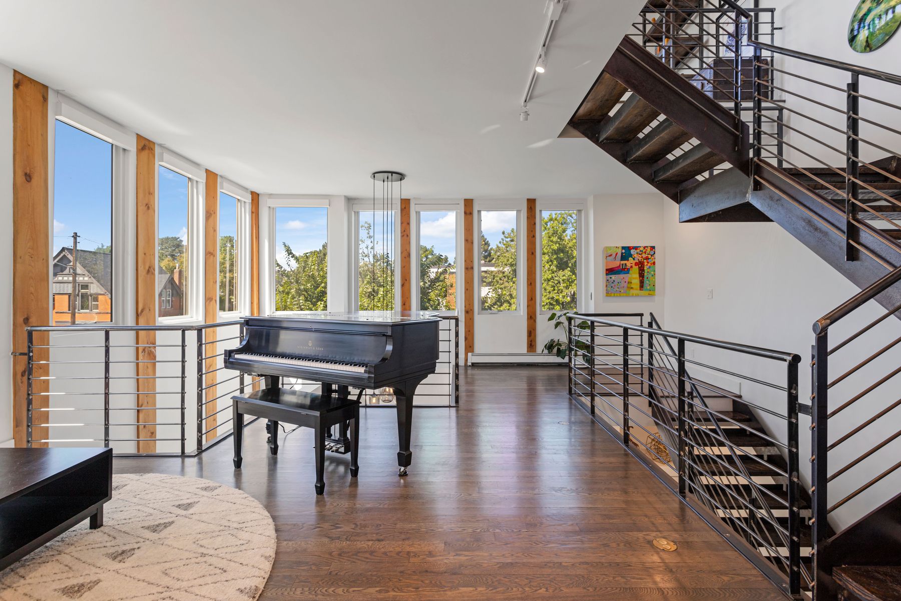 This is an interior shot of a modern living room featuring hardwood floors, large windows with wooden frames, and a black grand piano. A metal staircase with intricate railings adds an industrial touch, while a round rug softens the space. The room is bright and airy, creating an inviting atmosphere.