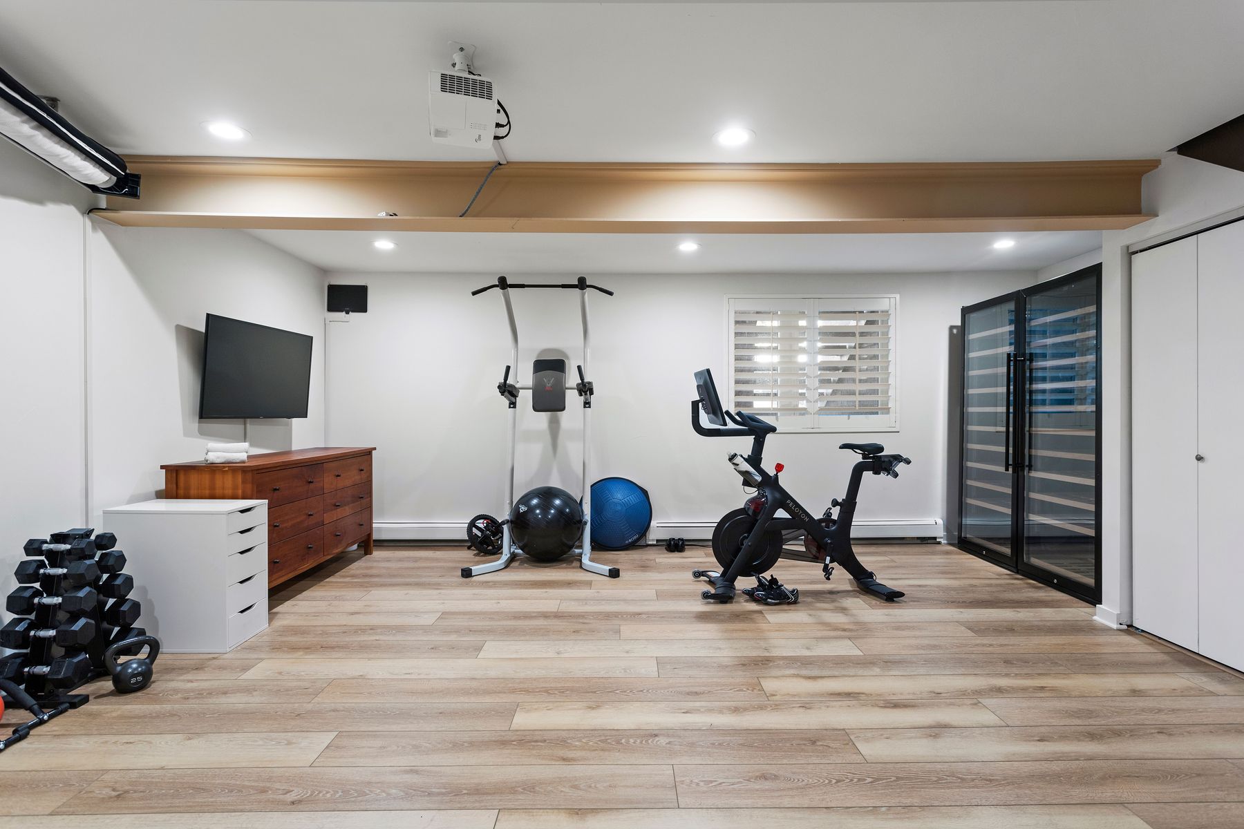 This is a well-equipped home gym featuring a Peloton bike, a pull-up station, dumbbells, and exercise balls. The room has light wood flooring, white walls, and a large window with blinds, creating a bright and inviting workout space. A TV and storage cabinets add functionality and convenience.