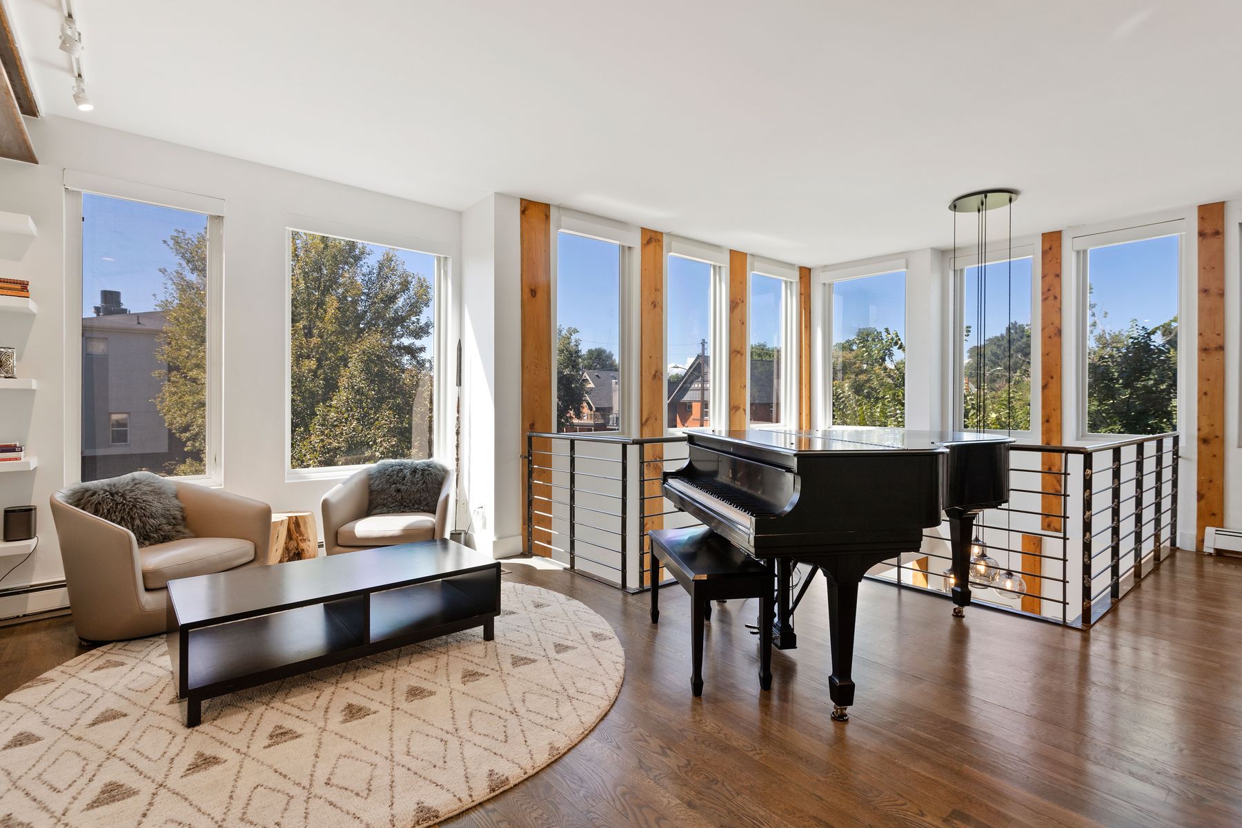 This is a bright and airy living room featuring hardwood floors, large windows with natural wood frames, and a black grand piano. The room is furnished with modern seating, a dark wood coffee table, and a round patterned rug, creating a sophisticated and inviting atmosphere. The space is well-lit and appears spacious, ideal for entertaining or relaxing.