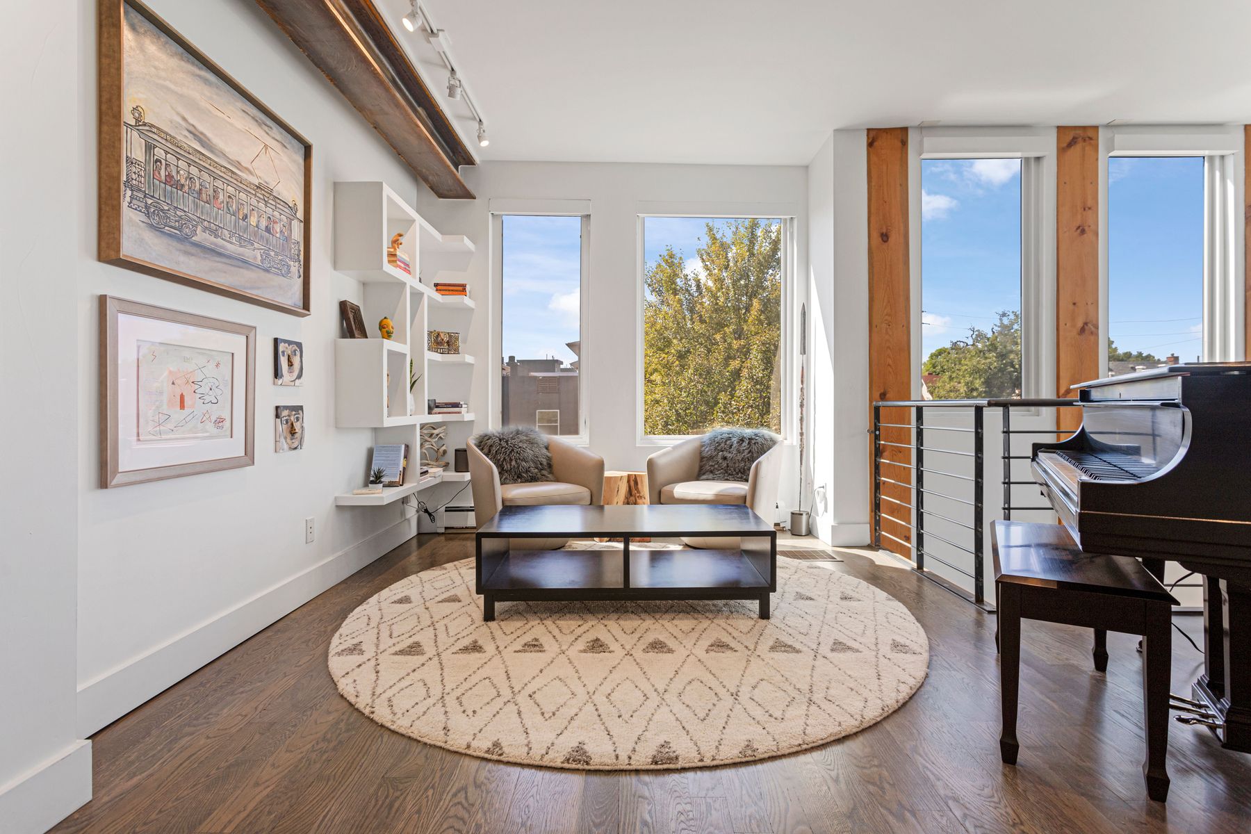 This is an inviting living room featuring hardwood floors and a neutral color palette. The room is furnished with two armchairs, a dark coffee table on a round patterned rug, and a black piano. Natural light floods the space through large windows, creating a bright and airy atmosphere.