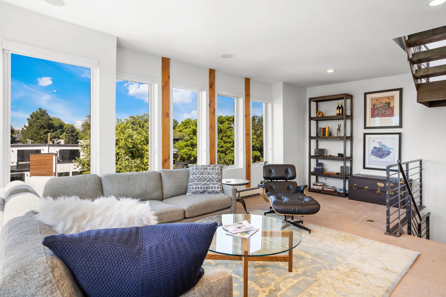 This is a bright and modern living room featuring a large sectional sofa with decorative pillows, a glass-top coffee table, and a black leather lounge chair. The room has large windows that provide ample natural light and views of the outdoors. A bookshelf and staircase add architectural interest to the space.