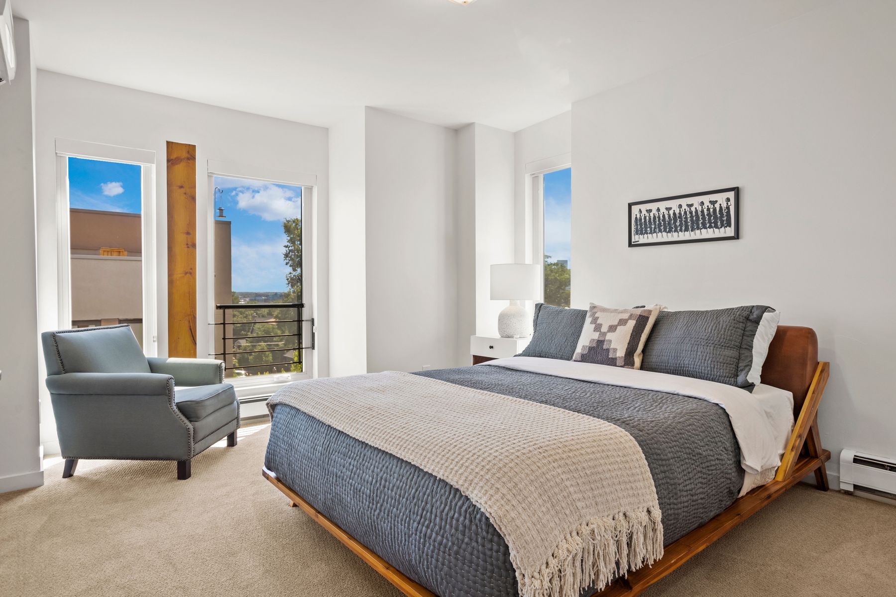 This is a bright and airy primary bedroom featuring a wooden bed frame with a gray quilted comforter and a textured throw blanket. The room has large windows that let in natural light, complemented by a comfortable armchair. A framed artwork hangs above the bed, adding a touch of sophistication to the minimalist design.