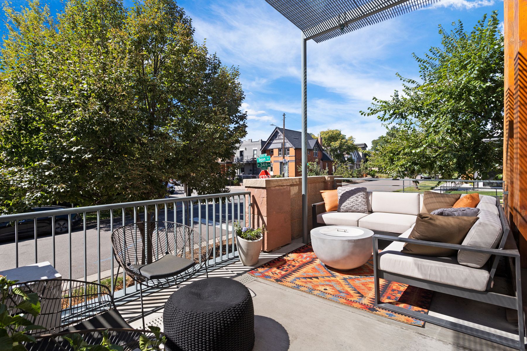This image showcases a stylish outdoor patio or balcony area, featuring modern furniture including a comfortable sectional sofa with throw pillows, wire chairs, and a round fire pit table. The space is decorated with an outdoor rug and potted plants, creating a cozy and inviting atmosphere. The view from the patio includes trees, a street, and nearby houses, suggesting an urban or suburban setting.