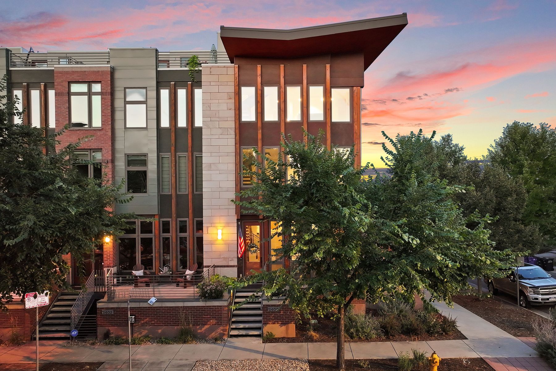 This is a front exterior view of a modern townhouse at dusk. The building features a mix of materials including brick, stone, and wood accents. The warm glow from the interior lights creates an inviting atmosphere, and the landscaping adds to the curb appeal.