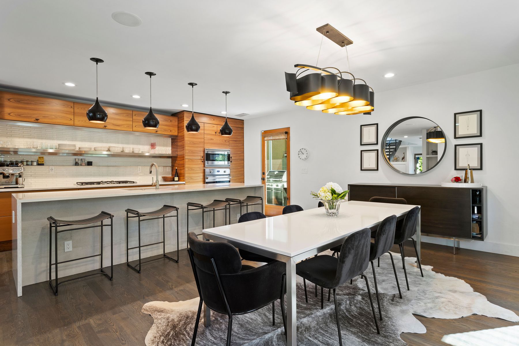 This interior shot showcases a modern dining room with a white table surrounded by black velvet chairs, set upon a cowhide rug. A contemporary chandelier hangs above the table, while a sideboard with a round mirror and framed artwork adorns the wall. The space is bright and stylish, blending modern and eclectic elements.
