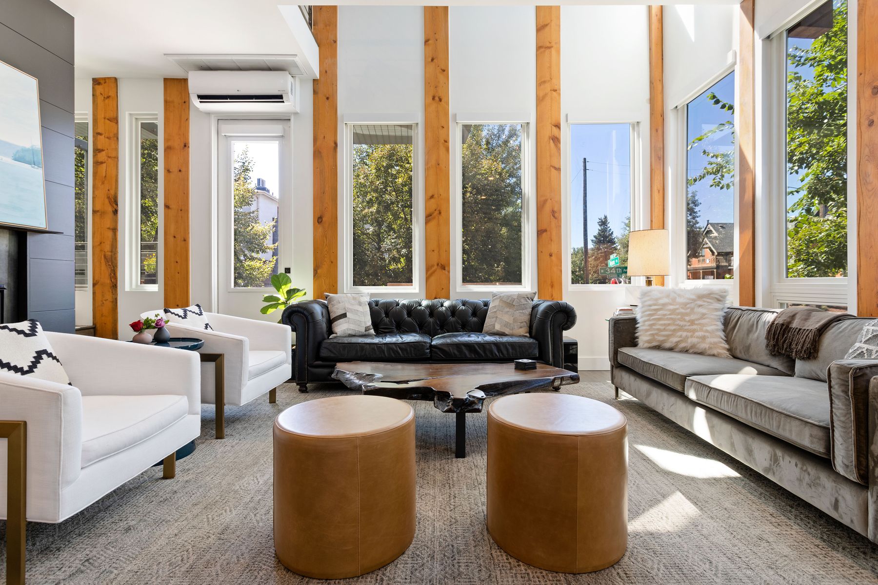 This is an interior shot of a modern living room featuring a black leather sofa, a gray sofa, and two white armchairs. The room is well-lit with natural light streaming through large windows framed by wooden beams. Two brown ottomans are in the center of the room, and a unique wooden coffee table sits between the sofas, adding to the room's contemporary and inviting atmosphere.