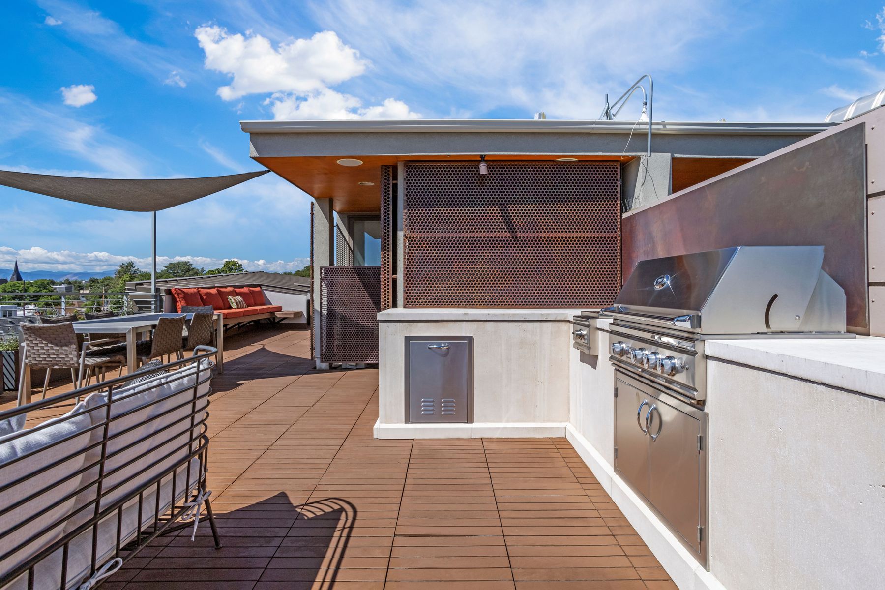This image showcases a stylish rooftop deck, perfect for outdoor entertaining. The deck features a built-in grill area with stainless steel appliances, comfortable seating arrangements with modern furniture, and a shade sail providing respite from the sun. The space is designed with a contemporary aesthetic, offering a luxurious outdoor living experience with a view.