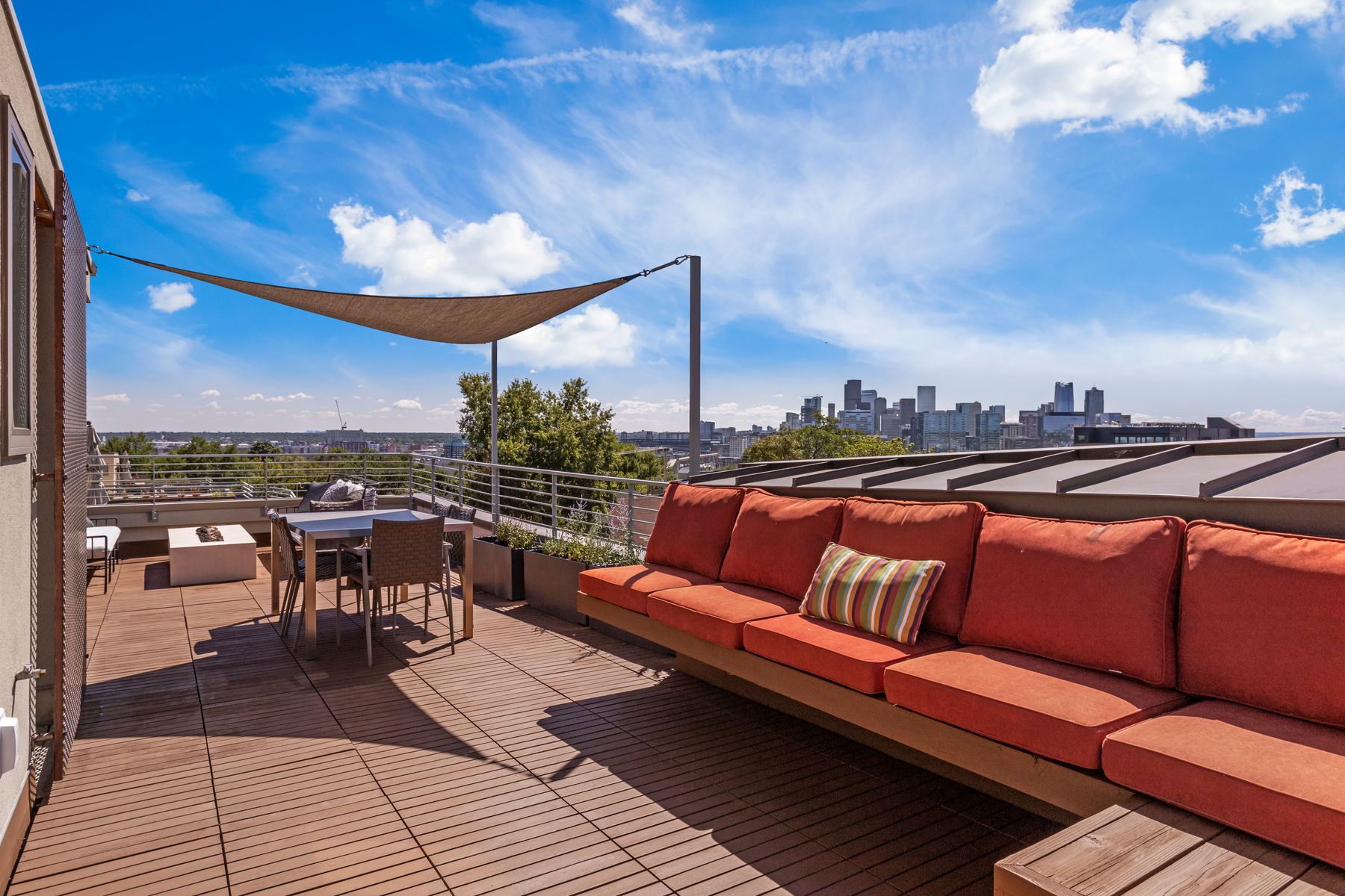 This image showcases a luxurious rooftop deck with stunning city views. The deck features a comfortable orange cushioned sofa with a striped accent pillow, a dining table with chairs, and a modern fire pit. The wooden deck flooring and shade sail add to the upscale ambiance, making it an ideal outdoor entertaining space.