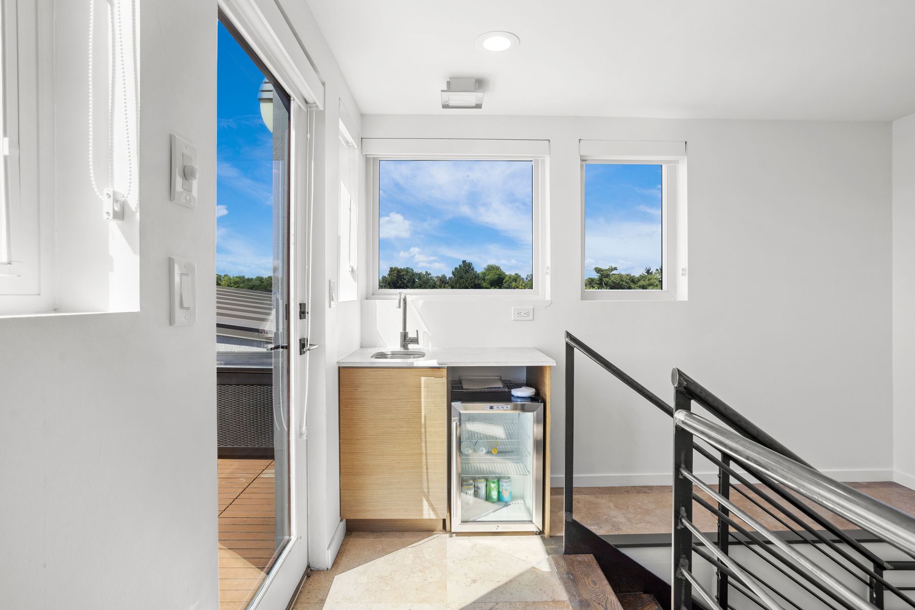 This interior shot showcases a bright hallway area with a small wet bar featuring a sink and a beverage refrigerator. Natural light floods the space through multiple windows, highlighting the clean, modern design and the staircase with metal railings. The flooring appears to be tile, adding to the contemporary aesthetic.
