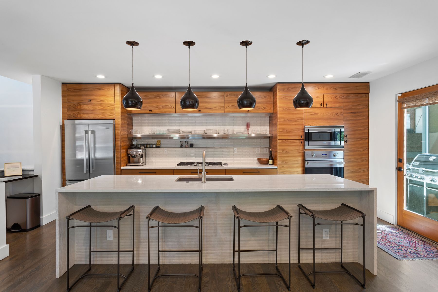 This is a well-lit, modern kitchen featuring wood-grain cabinetry and a large central island with a white countertop and seating for four. Stainless steel appliances, including a refrigerator and built-in oven/microwave, add to the contemporary feel. The kitchen is illuminated by pendant lights hanging above the island, and a door to the outside is visible on the right.