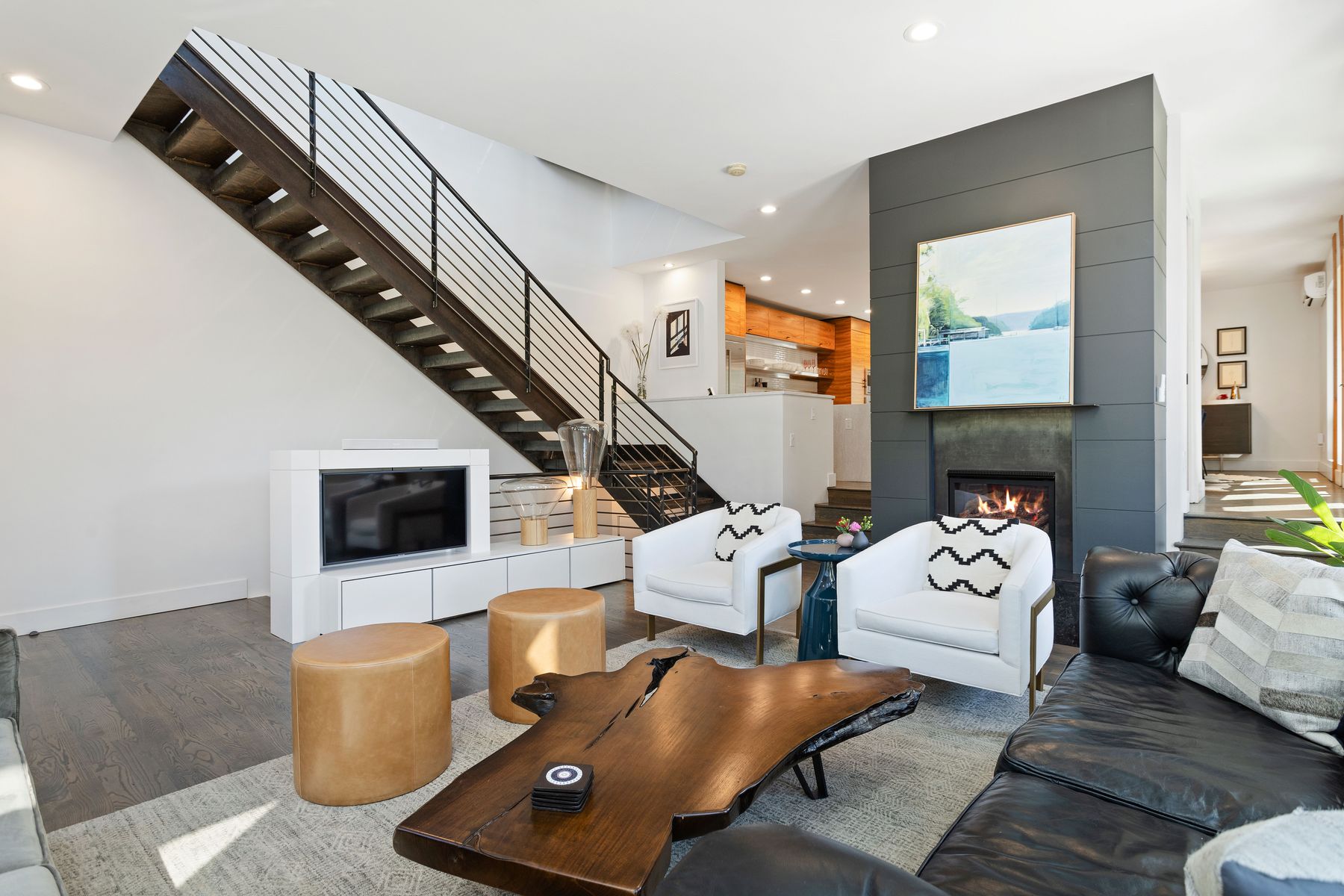 This is a stylish living room featuring a modern staircase with metal railings, a white media console with a flat-screen TV, and a unique live-edge coffee table. Two white armchairs with patterned pillows face a dark leather sofa, and a fireplace with a gray surround adds a focal point. The room is well-lit and has a contemporary design.