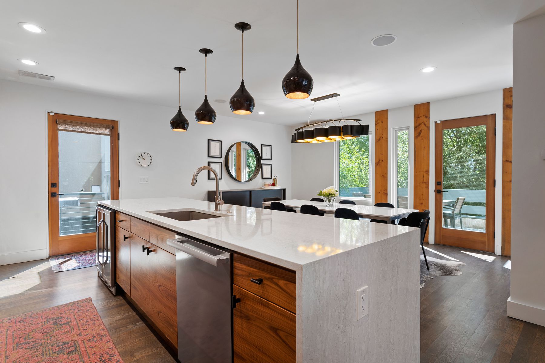 This is a bright and modern kitchen featuring a large island with a white countertop and wood cabinetry. Stainless steel appliances are integrated seamlessly into the design. Pendant lights hang above the island, and a dining area is visible in the background, creating an open and inviting space.