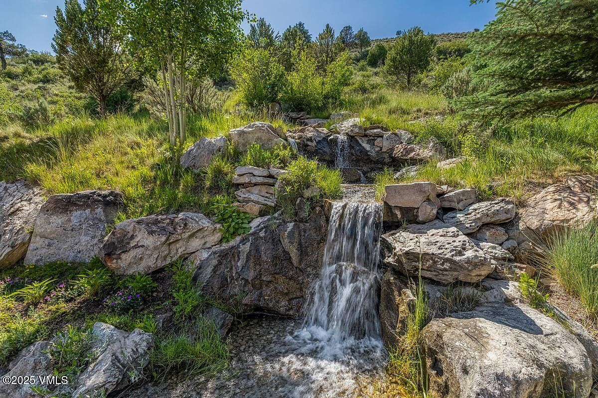 This image showcases a beautifully landscaped yard featuring a multi-tiered rock waterfall. Lush greenery and mature trees surround the water feature, creating a serene and natural ambiance. The scene suggests a tranquil outdoor space perfect for relaxation and enjoying nature.