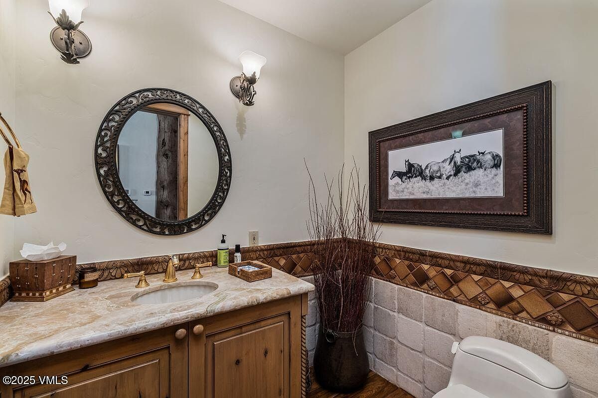 This is a well-appointed guest bathroom featuring a rustic vanity with a marble countertop and a round, ornate mirror. The walls are adorned with decorative tile and a framed picture of horses, adding a touch of Western charm. The room is illuminated by sconce lighting, creating a warm and inviting atmosphere.