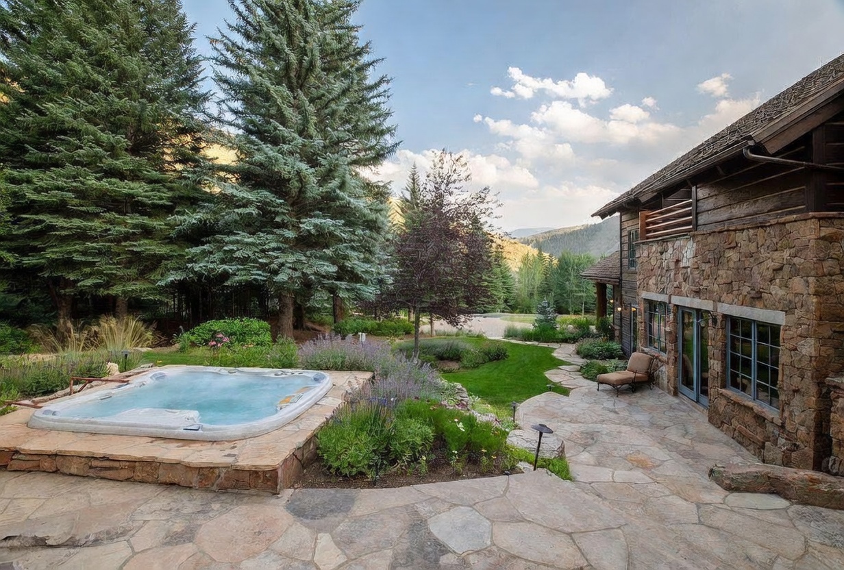 This image showcases a beautifully landscaped backyard featuring a stone patio, a built-in hot tub, and lush greenery. The stone facade of the house complements the natural surroundings, creating a serene and inviting outdoor space. Mature trees provide privacy and shade, enhancing the property's appeal.